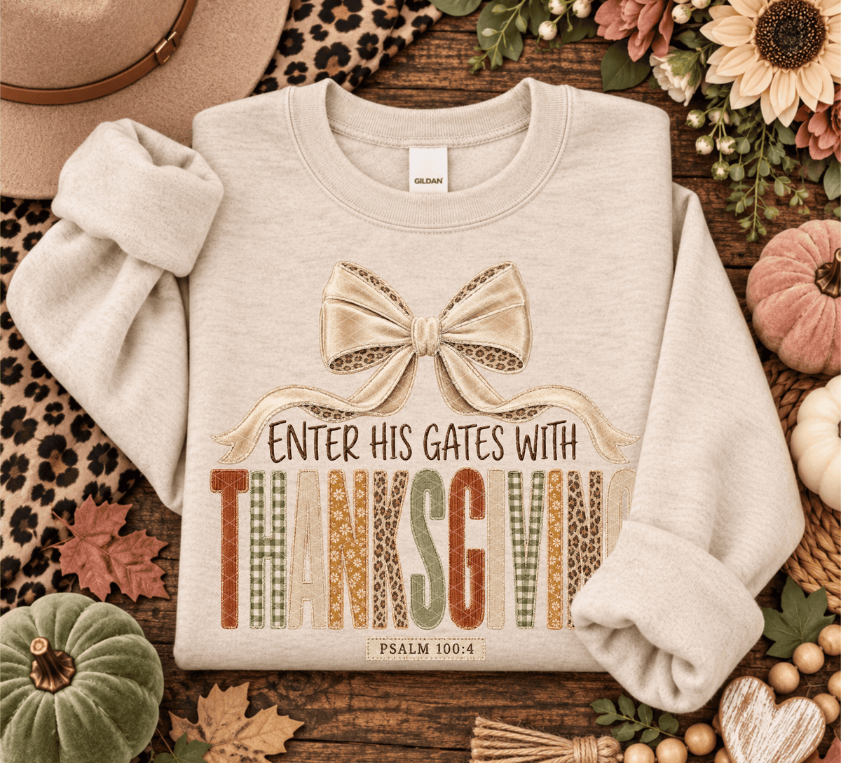 A light beige crewneck sweatshirt laid flat on a rustic wood surface, surrounded by fall-themed props including pumpkins, leaves, flowers, and a hat. The sweatshirt features a large bow graphic and the text 'ENTER HIS GATES WITH THANKSGIVING' and 'PSALM 100:4' printed on the front.