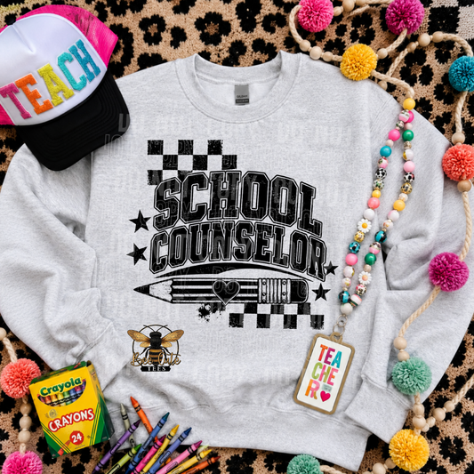 A light heather gray crewneck sweatshirt with a black 'SCHOOL COUNSELOR' graphic, styled in a flat lay with a 'TEACH' baseball cap, crayons, a box of Crayola crayons, and colorful pom-poms on a leopard print background.