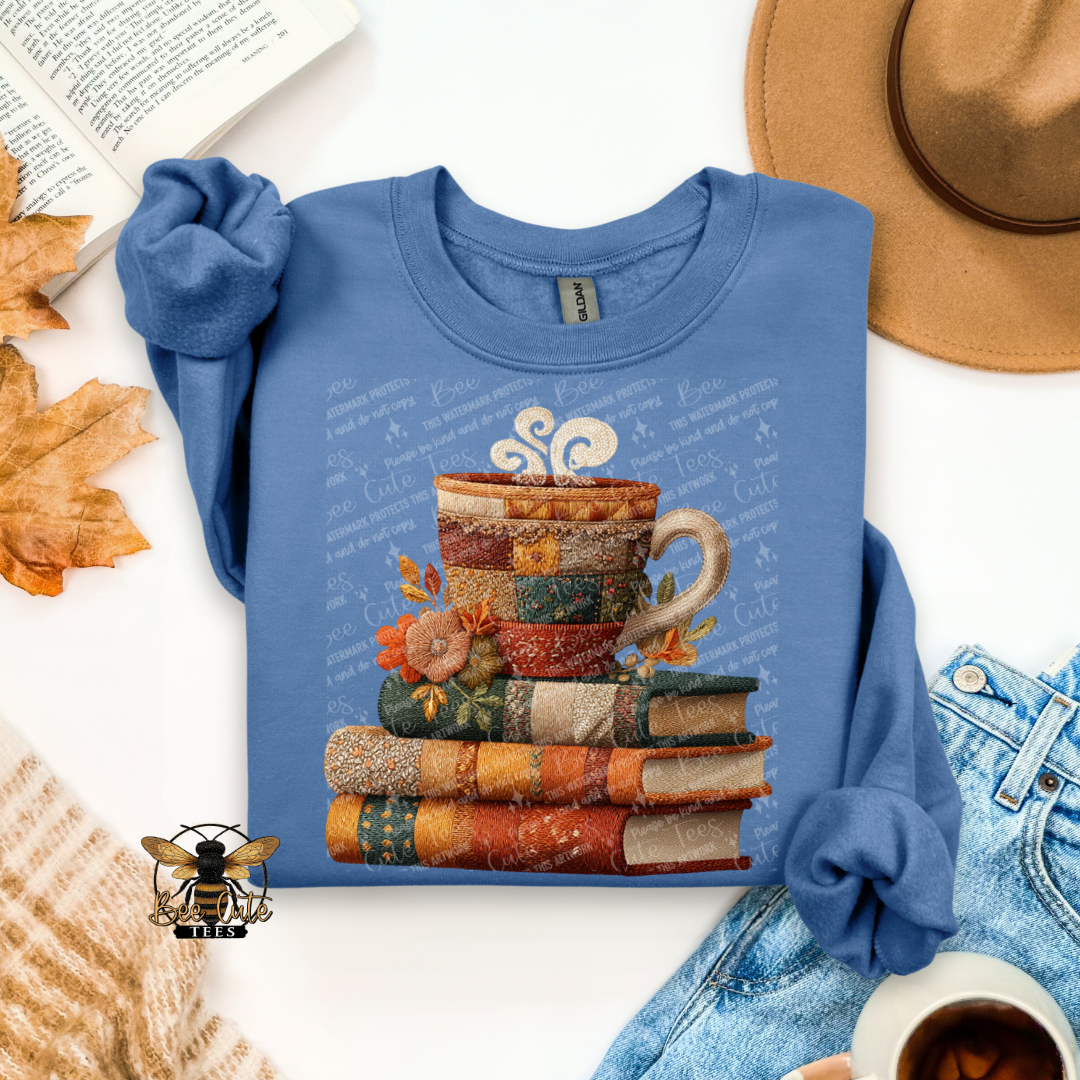 A folded blue long-sleeve shirt with rolled cuffs, featuring a graphic of a patchwork coffee mug resting on a stack of books. The shirt is styled in a flat-lay arrangement with a tan hat, blue jeans, a book, autumn leaves, a cup of coffee, and a cozy knit sweater on a white background.