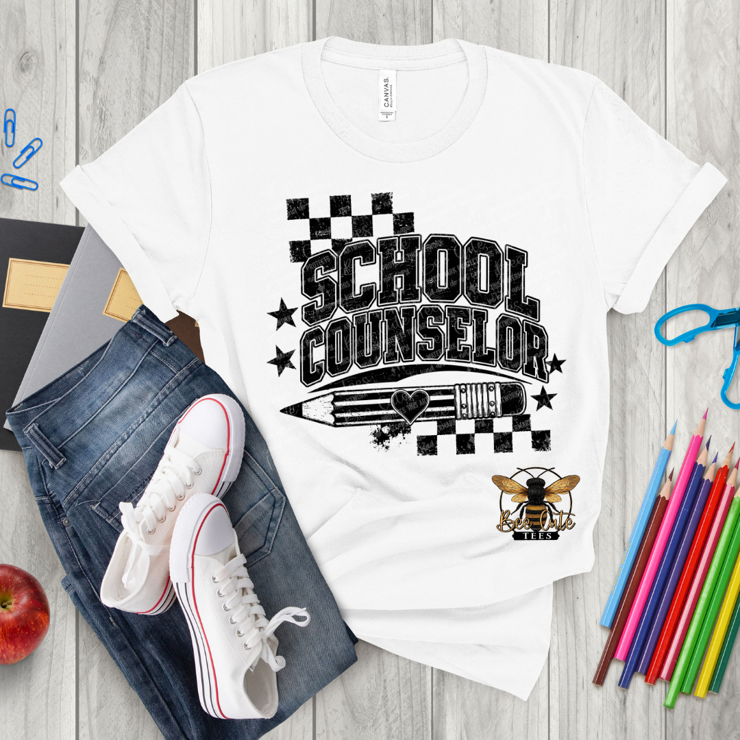 A white graphic t-shirt featuring a black 'SCHOOL COUNSELOR' design with a pencil, stars, and a bee mascot, styled in a flat lay with jeans, sneakers, colored pencils, and an apple on a light wood background.