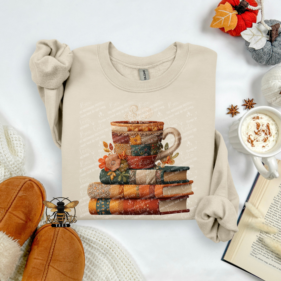 A beige long-sleeve crewneck sweatshirt with a graphic of stacked books topped with a patterned mug, styled in a cozy flat-lay with slippers, a mug of hot cocoa, an open book, and autumn decorations.