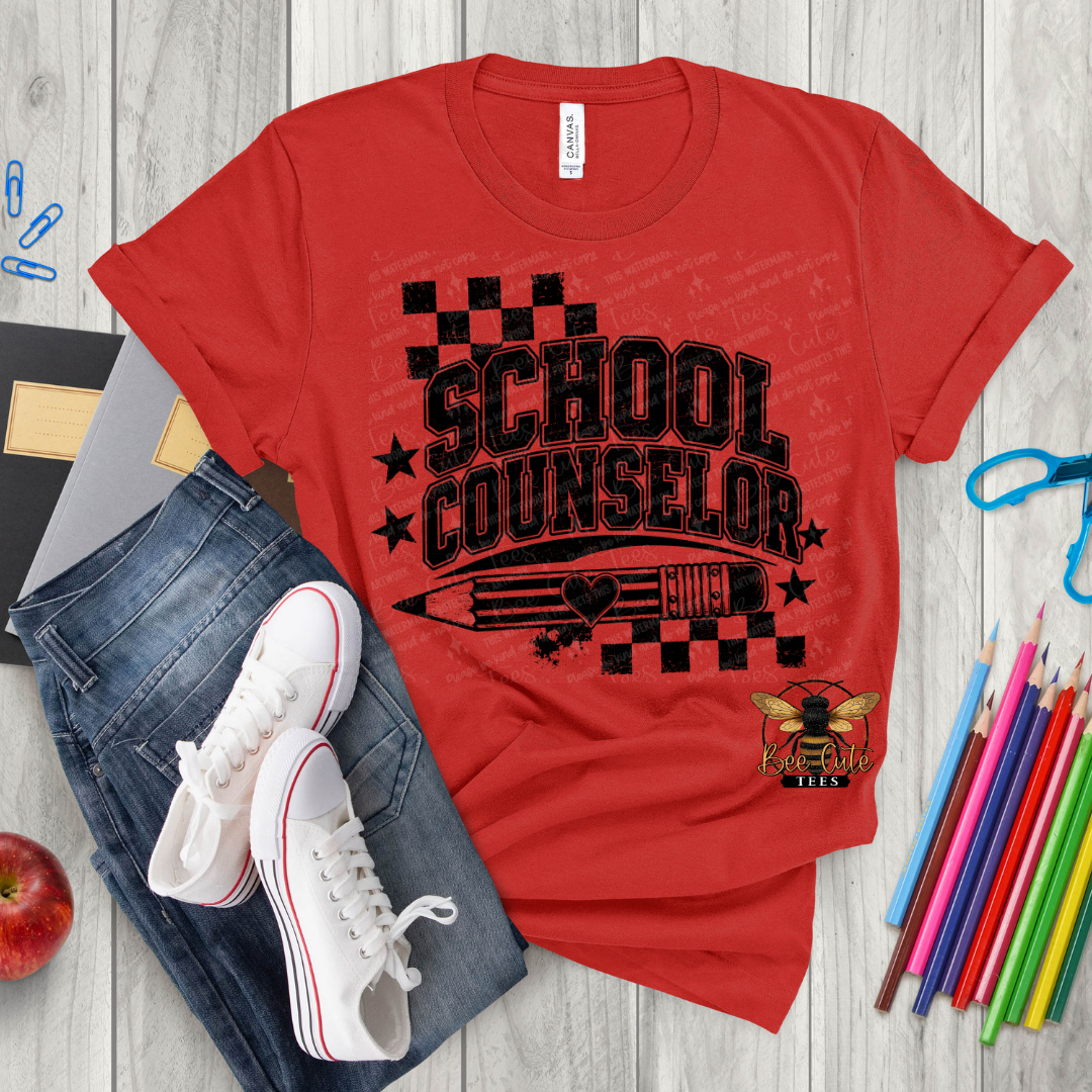 A red graphic T-shirt laid flat on a light wood surface, styled with blue jeans, white sneakers, colored pencils, a apple, and small office props. The shirt has a large black 'SCHOOL COUNSELOR' text design with checkered flag elements, stars, a pencil graphic, and a bee emblem near the lower right.