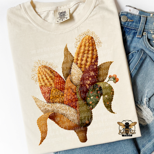 Folded ivory Comfort Colors t-shirt featuring a vintage cottage-style fall harvest design of colorful corn with faux embroidery and patchwork textures in shades of mustard yellow, burnt orange, rust, cream, and olive green. Styled with distressed blue jeans and a Bee Cute Tees watermark.