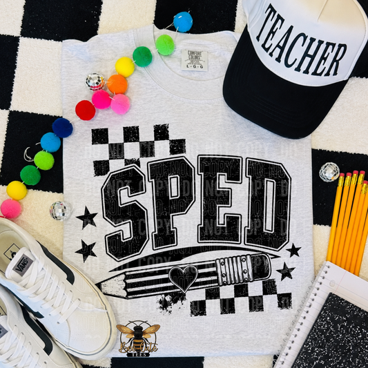 A flat-lay of a white graphic t-shirt featuring the word 'SPED' in distressed black varsity letters with a pencil and stars. The shirt is styled with a black and white checkered rug, a black and white 'TEACHER' cap, colorful pom-pom beads, a white sneaker, yellow pencils, and a notebook.