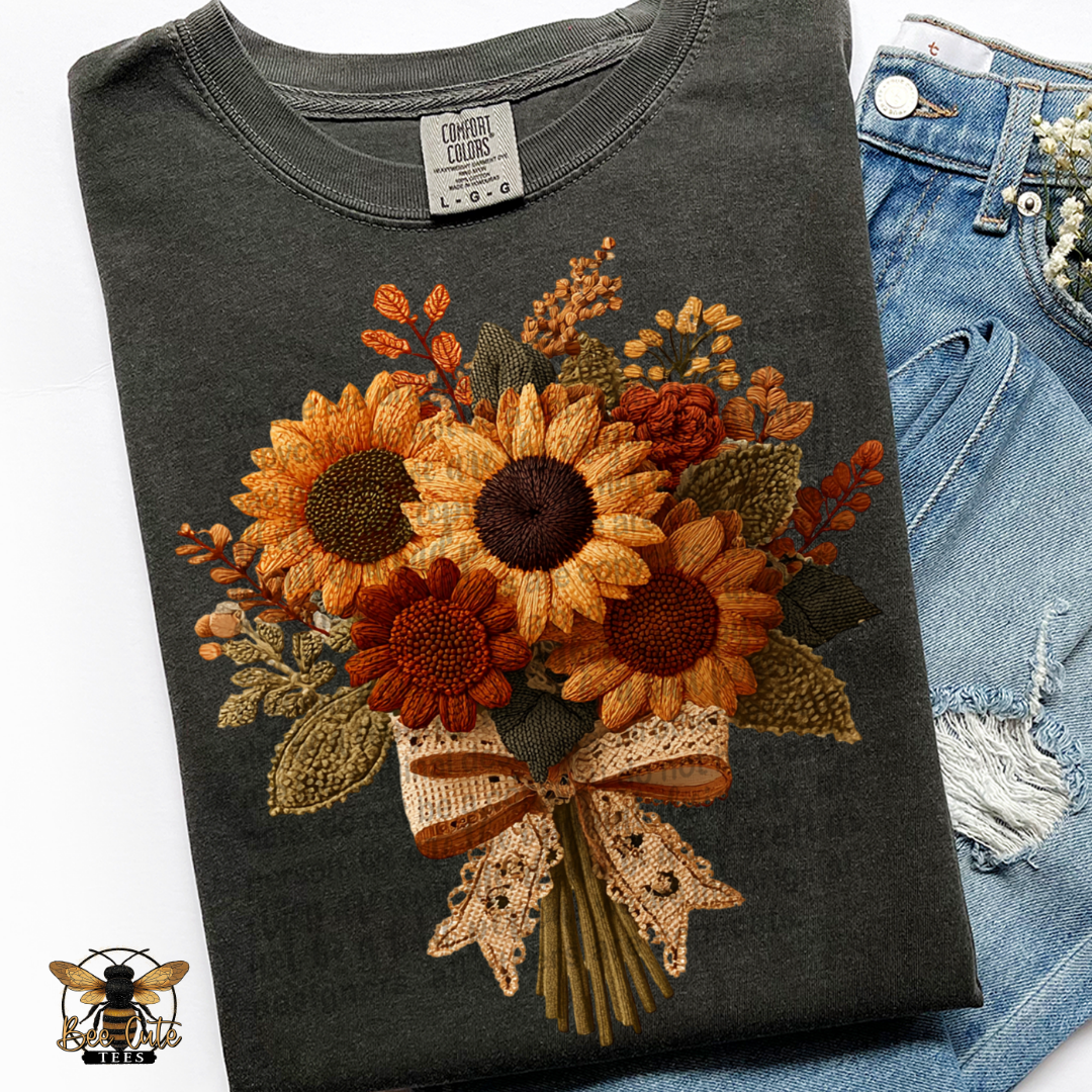 Folded charcoal gray Comfort Colors t-shirt featuring a vintage cottage-style sunflower bouquet with faux embroidery textures. Golden yellow, burnt orange, rust, and olive green flowers and foliage are tied with a cream lace bow. Styled with distressed blue jeans and a Bee Cute Tees watermark.