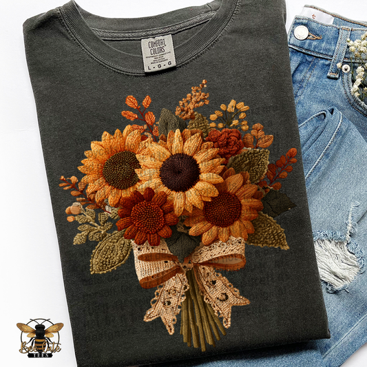 Folded charcoal gray Comfort Colors t-shirt featuring a vintage cottage-style sunflower bouquet with faux embroidery textures. Golden yellow, burnt orange, rust, and olive green flowers and foliage are tied with a cream lace bow. Styled with distressed blue jeans and a Bee Cute Tees watermark.