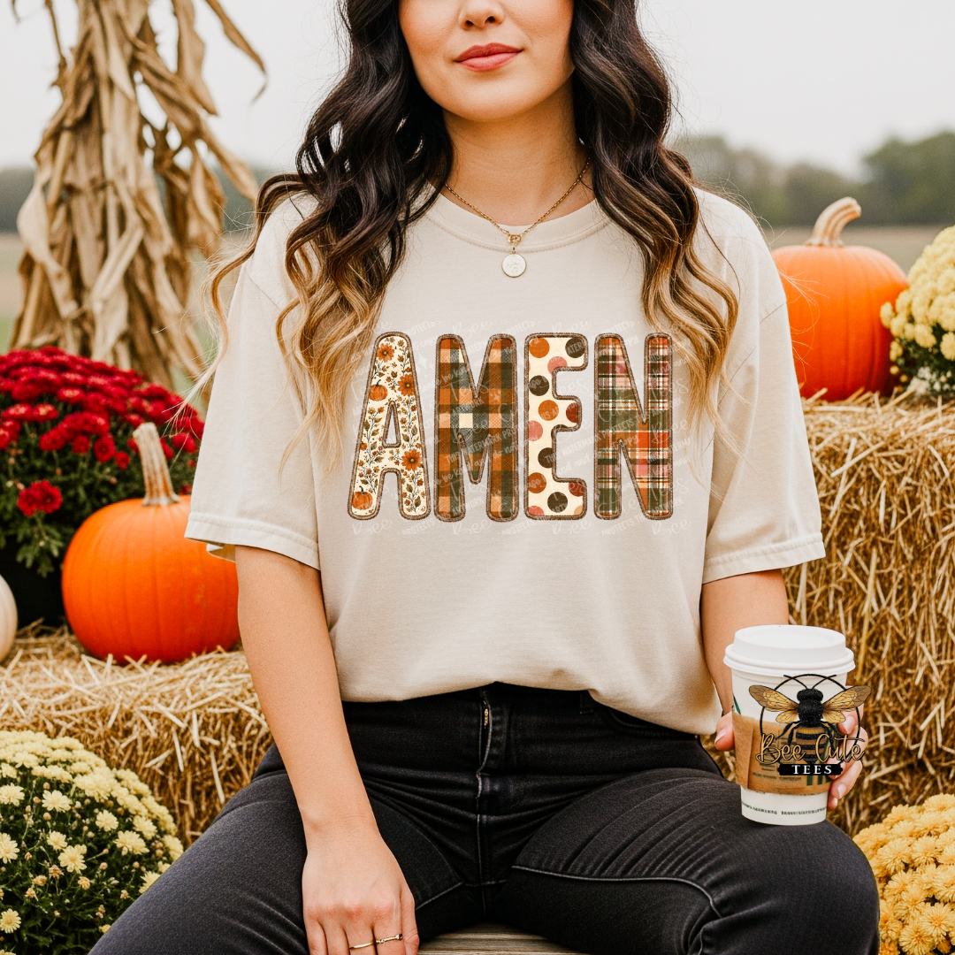 A woman wearing a beige graphic t-shirt with the word 'AMEN' in autumn-themed plaid and leopard print patterns, sitting in a fall-themed setting with pumpkins, hay bales, and a coffee cup.