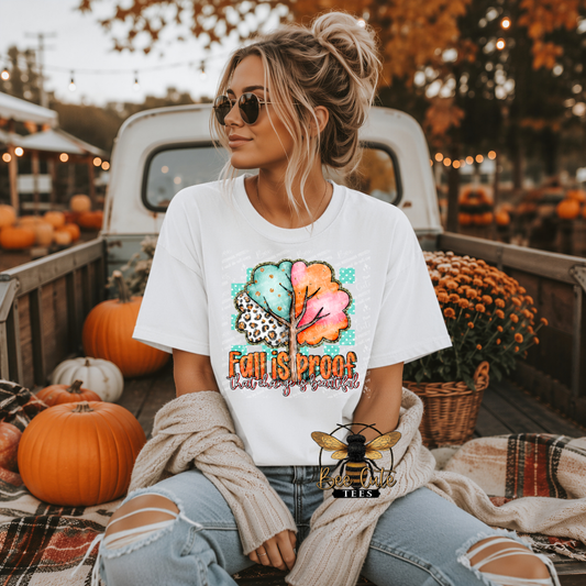 A woman wearing a white graphic t-shirt with a colorful autumn leaf design and the text 'Fall is Proof' sits in the bed of a pickup truck decorated for fall. She is wearing a beige knit sweater draped over her shoulders and ripped light-wash jeans.