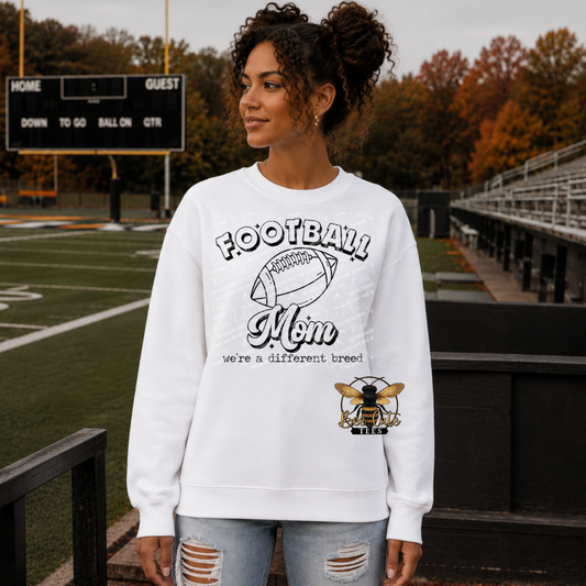 A woman wearing a white crewneck sweatshirt with a 'Football Mom' graphic and bee logo, standing in front of a football field and stadium.