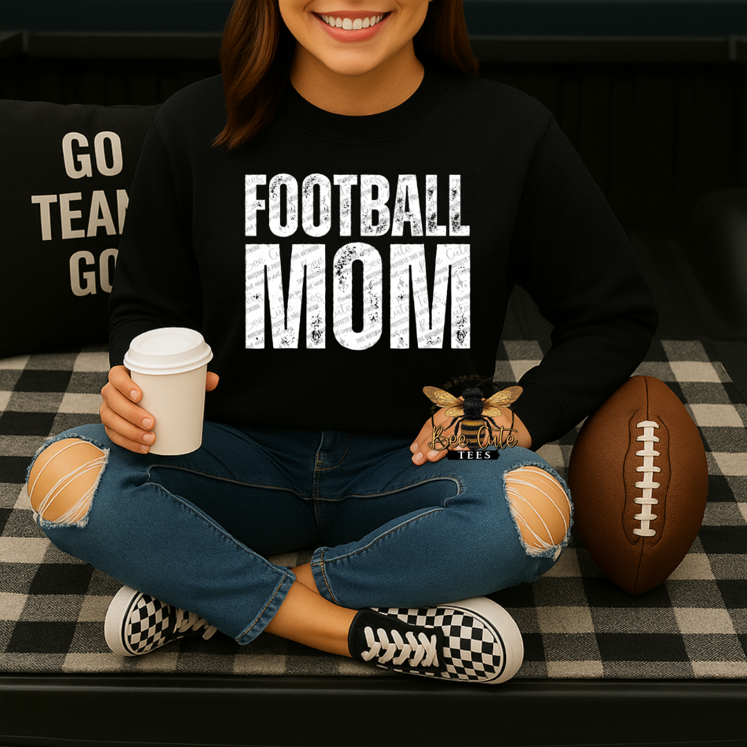 A woman sitting cross-legged on a black-and-white checkered couch, wearing a black sweatshirt with 'FOOTBALL MOM' printed in distressed white letters. She holds a coffee cup and a small bee ornament, with a football beside her.