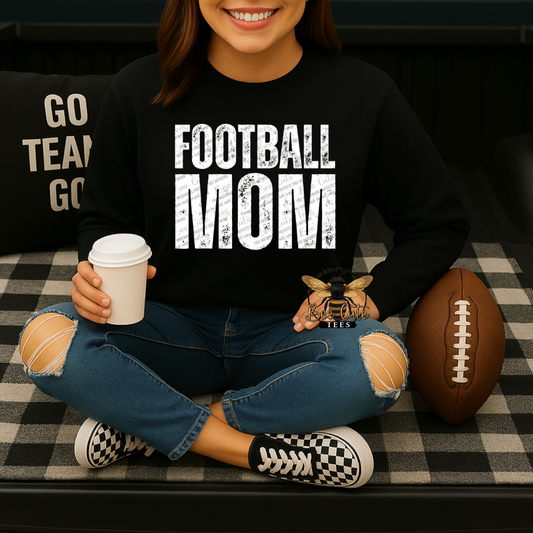 A woman sitting cross-legged on a black-and-white checkered couch, wearing a black sweatshirt with 'FOOTBALL MOM' printed in distressed white letters. She holds a coffee cup and a small bee ornament, with a football beside her.