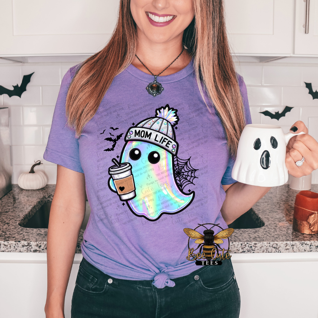A woman wearing a purple graphic t-shirt with a cartoon ghost wearing a 'Mom Life' beanie and holding a coffee cup. She is holding a white ghost-shaped mug in a kitchen setting.