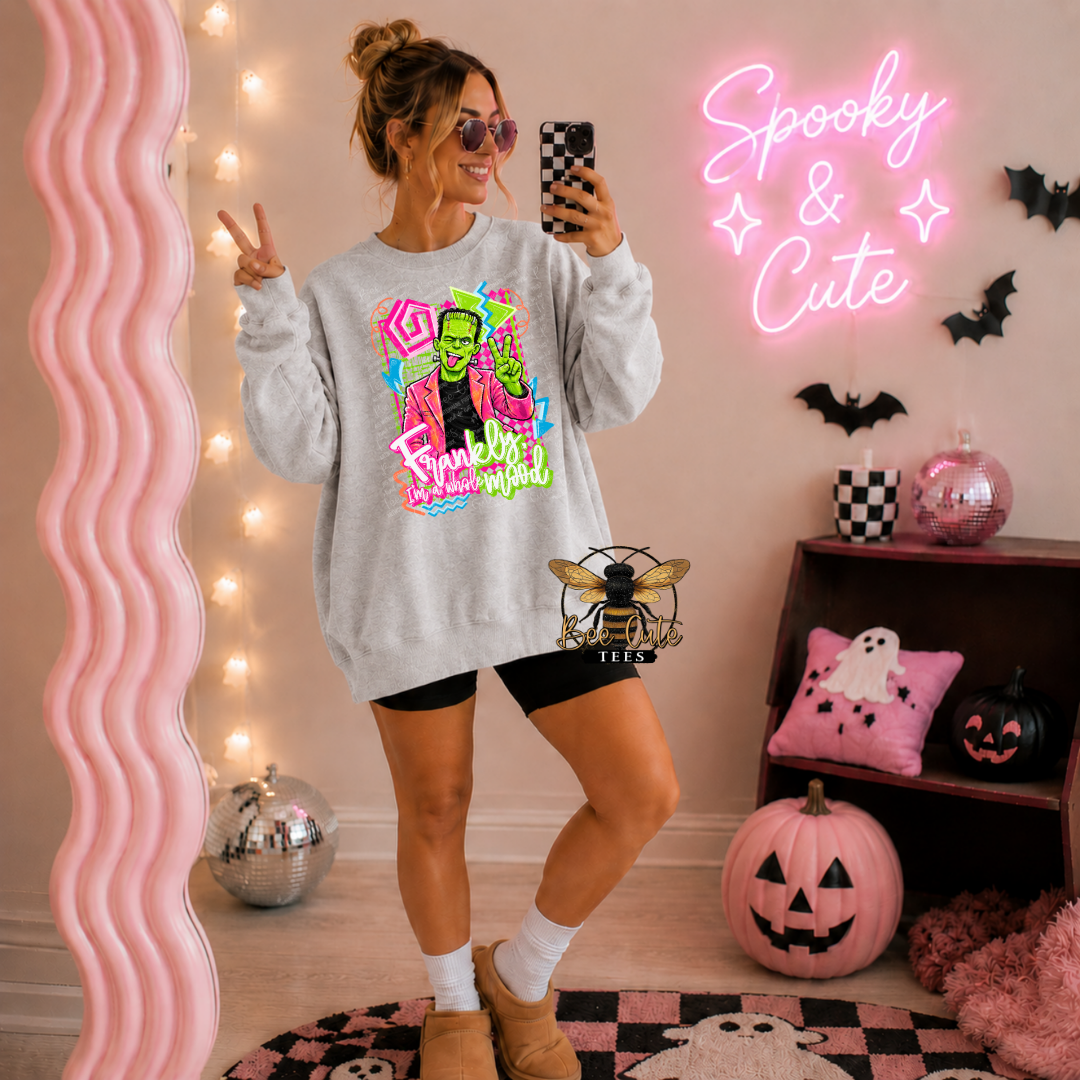 A woman takes a mirror selfie wearing a light gray crewneck sweatshirt with a colorful 'Frankenstein's Monster' graphic. The room is decorated for Halloween with a pink wavy mirror, a pink neon sign reading 'Spooky & Cute', pumpkins, and a shelf with ghost and pumpkin pillows.