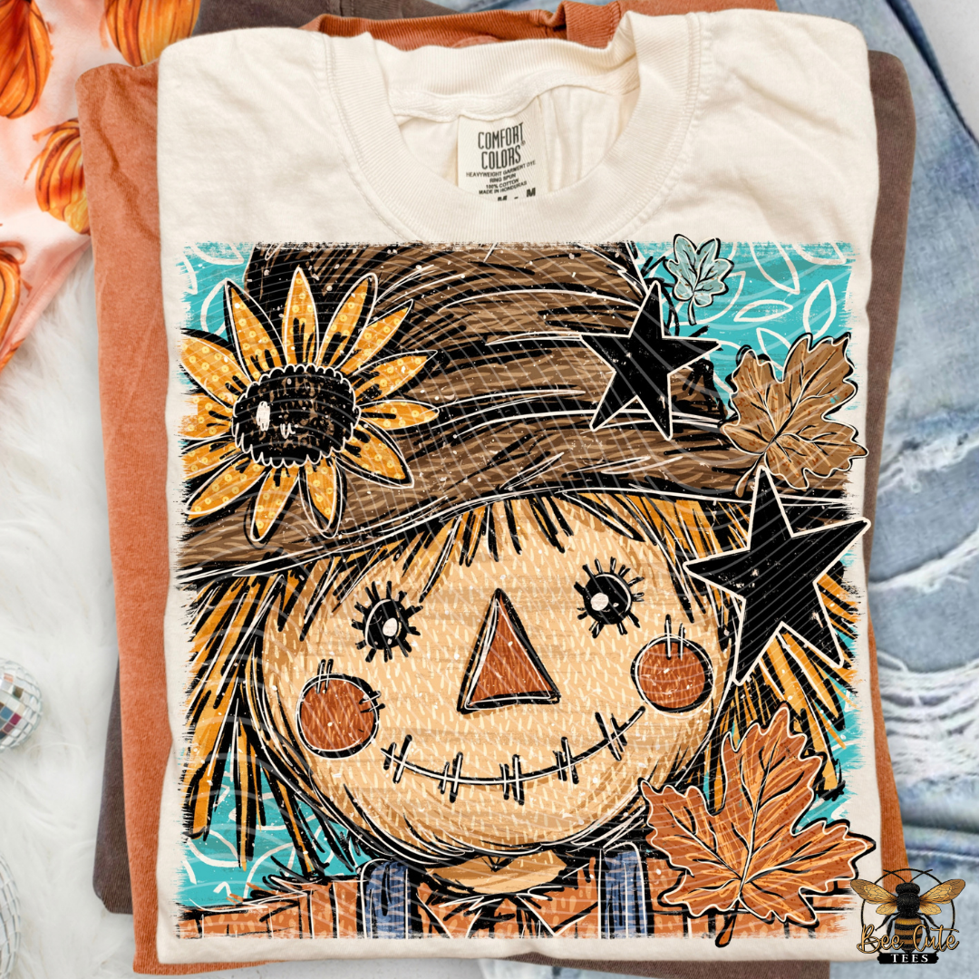 A folded cream-colored t-shirt featuring a colorful graphic print of a scarecrow with a sunflower, autumn leaves, stars, and a blue patterned background. The shirt is styled on top of another orange shirt with a rustic flat-lay background.