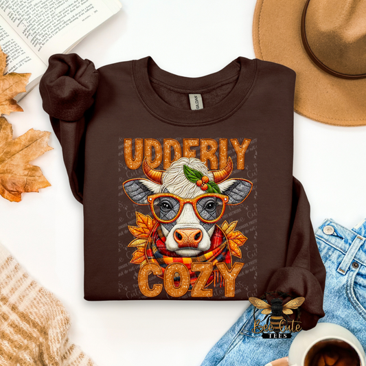 A folded dark brown long-sleeve t-shirt with a graphic print of a cow wearing orange glasses and a red scarf, surrounded by autumn leaves and fall colors. The text 'UDDERLY COZY' is printed above and below the cow.