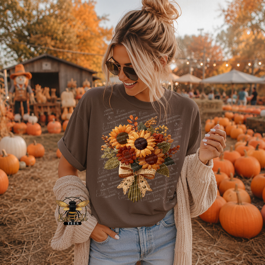 Woman wearing a brown graphic t-shirt featuring a vintage fall sunflower bouquet with faux embroidered flowers in warm autumn shades and a delicate lace bow. Styled with jeans, sunglasses, and a cream cardigan at a festive pumpkin patch. Bee Cute Tees watermark visible.