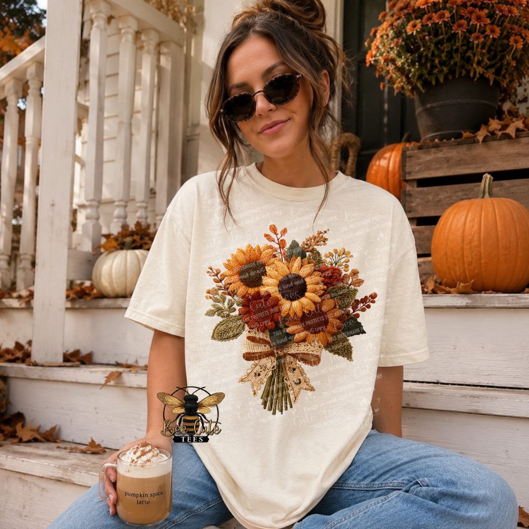 Woman wearing an ivory graphic t-shirt featuring a cottage-style fall bouquet of sunflowers, rust-colored flowers, and greenery with faux embroidery textures and a lace ribbon bow. Seated on autumn-decorated porch steps holding a pumpkin spice latte, with a Bee Cute Tees watermark.