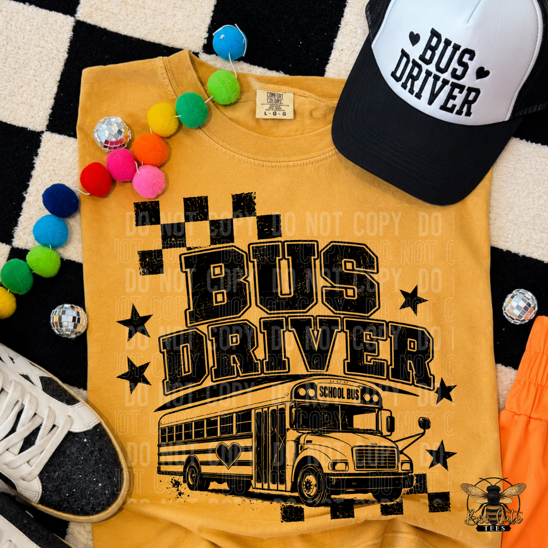 A yellow graphic t-shirt featuring a black 'BUS DRIVER' text design with a school bus illustration, styled in a flat lay with a black and white trucker hat, colorful pom-poms, a sneaker, and a checkered rug.