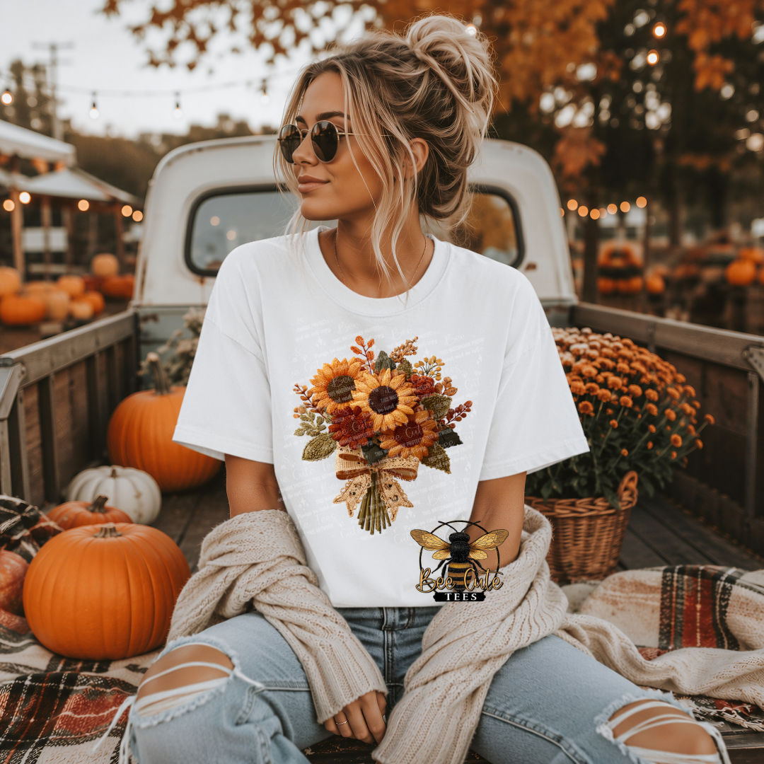 Woman wearing a white graphic t-shirt featuring a vintage sunflower bouquet with faux embroidered golden yellow and rust flowers, olive green leaves, and a cream lace bow. Styled with distressed jeans and a cozy cardigan while sitting in a vintage pickup truck surrounded by pumpkins and fall flowers. Bee Cute Tees watermark visible.