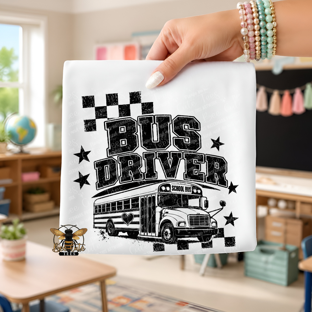 A hand holds a white folded t-shirt or small flag featuring a black distressed 'BUS DRIVER' graphic with a school bus illustration and stars, set against a blurred classroom background.