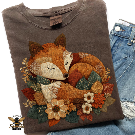 Folded vintage brown Comfort Colors t-shirt featuring a sleeping fox curled among autumn flowers and leaves. Cottage-style patchwork design with faux embroidery textures in burnt orange, rust, cream, mustard, and forest green. Styled with distressed blue jeans and a Bee Cute Tees watermark.