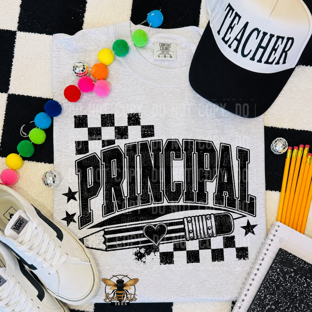 A white graphic t-shirt featuring the word 'PRINCIPAL' in distressed black block letters over a pencil and ruler design with checkerboard accents. The shirt is styled in a flat lay with a black and white checkered rug, a black and white 'TEACHER' cap, colorful pom-pom beads, a white sneaker, yellow pencils, and a notebook.