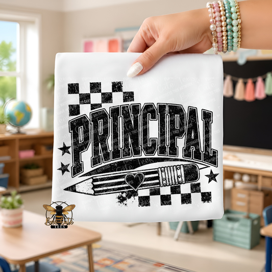 A hand holds up a white t-shirt featuring a black graphic design that reads 'PRINCIPAL' with a pencil, stars, and checkered flags. The background shows a blurred classroom interior.
