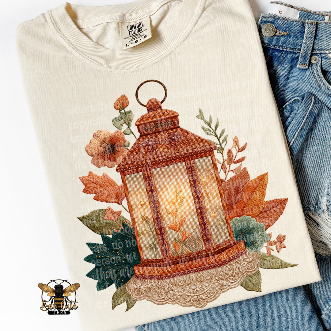 Folded ivory Comfort Colors t-shirt featuring a vintage cottage-style lantern glowing with warm light, surrounded by autumn leaves, flowers, and greenery. Faux embroidery textures in burnt orange, rust, cream, and sage green with delicate lace detailing. Styled with distressed blue jeans and a Bee Cute Tees watermark.