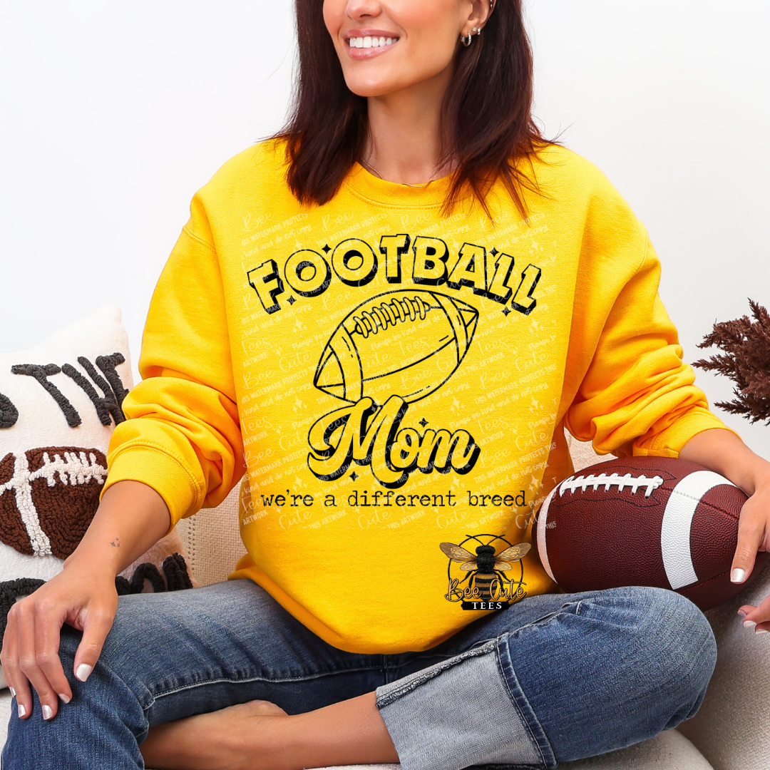 A woman wearing a bright yellow sweatshirt with a 'Football Mom' graphic sits on a couch holding a football. The sweatshirt features a distressed texture and a bee logo at the bottom right.