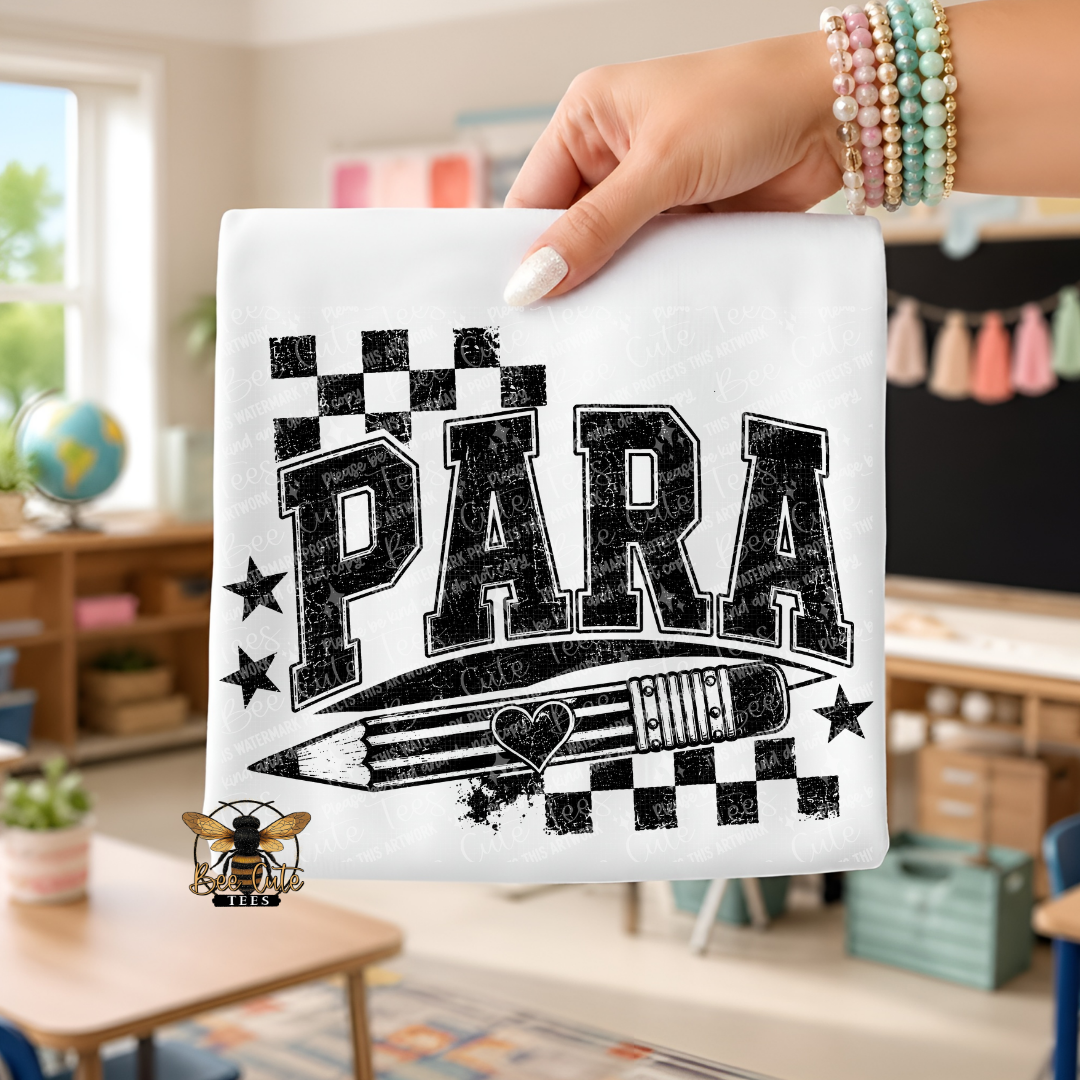 A hand holds up a white folded t-shirt featuring a black distressed graphic that reads 'PARA' with stars, a pencil, and checkerboard accents. The background shows a blurred classroom or home interior with shelves, a globe, and decor.