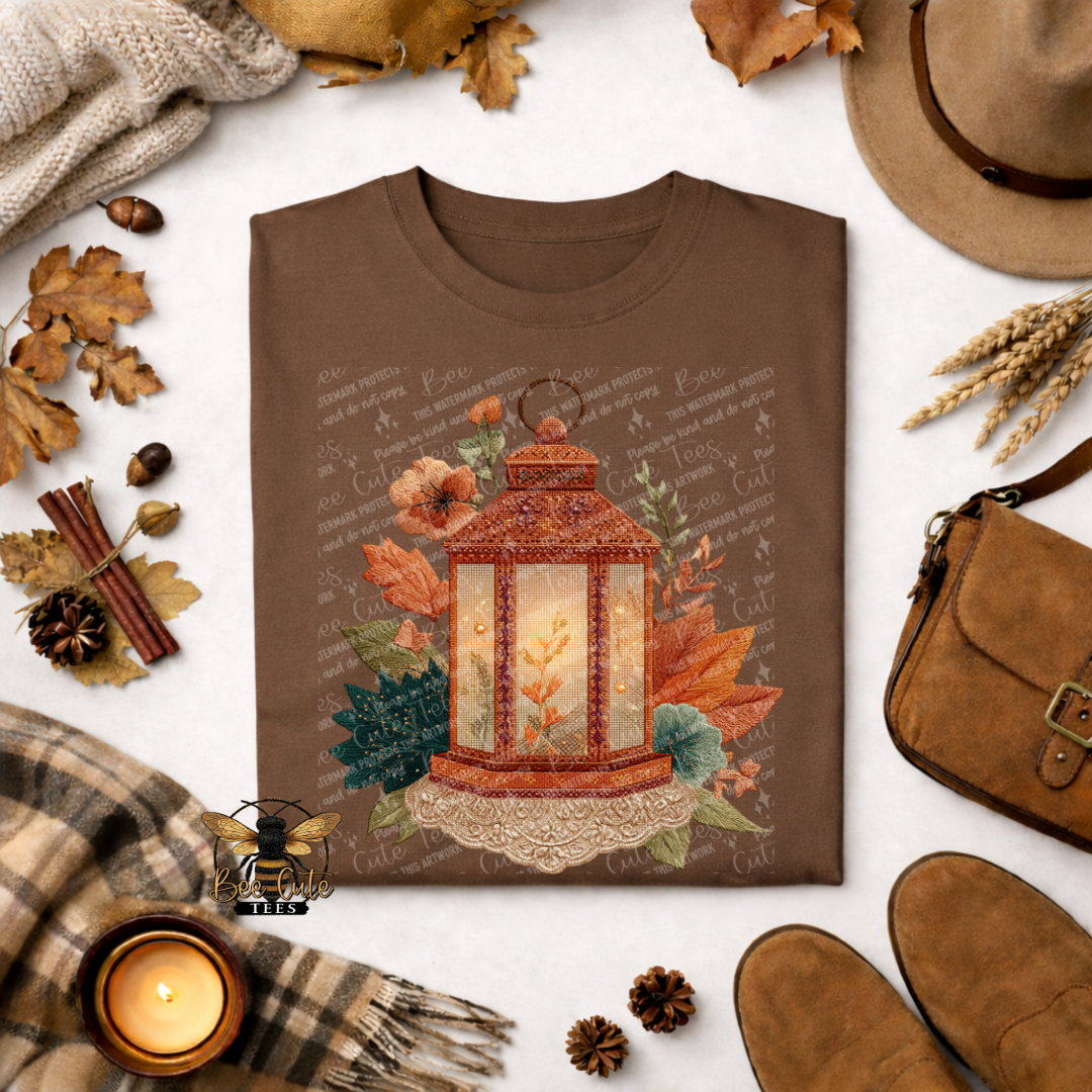 Folded brown t-shirt featuring a rustic lantern glowing with warm golden light, surrounded by autumn leaves, florals, and greenery in a vintage faux embroidery style. Styled with a plaid scarf, candle, pinecones, dried wheat, and fall accessories. Bee Cute Tees watermark visible.
