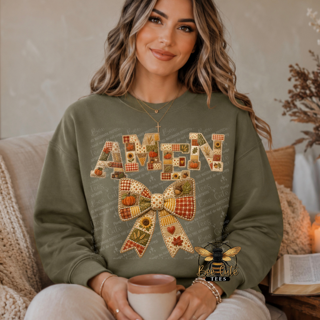 A woman wearing an olive green crewneck sweatshirt with a large autumn-themed 'AMEN' bow graphic, sitting on a couch and holding a mug.