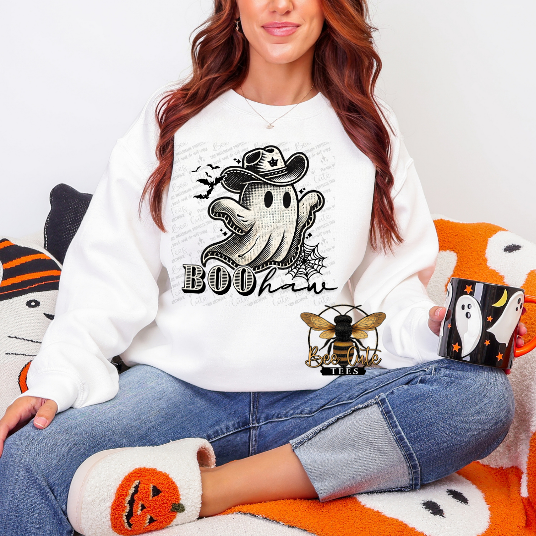 A woman wearing a white crewneck sweatshirt with a black and white ghost graphic that says 'BOOHaw' sits on a cozy orange and white blanket. She is holding a black mug with a ghost design and wearing white slippers with orange pumpkin details.
