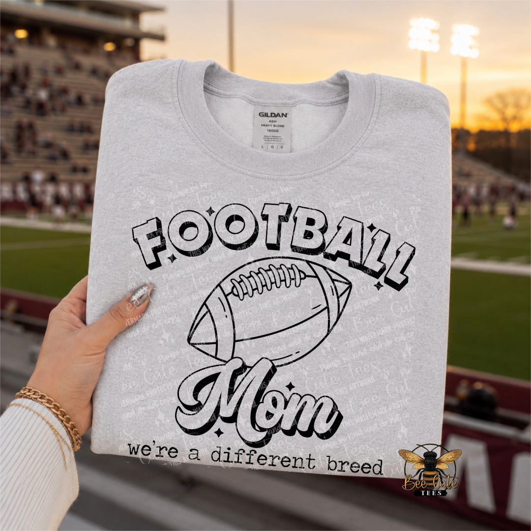 A folded light gray Gildan sweatshirt held by a hand, featuring a black graphic that reads 'FOOTBALL Mom' with a football illustration and the phrase 'we're a different breed.' The background shows a blurred football stadium at sunset.