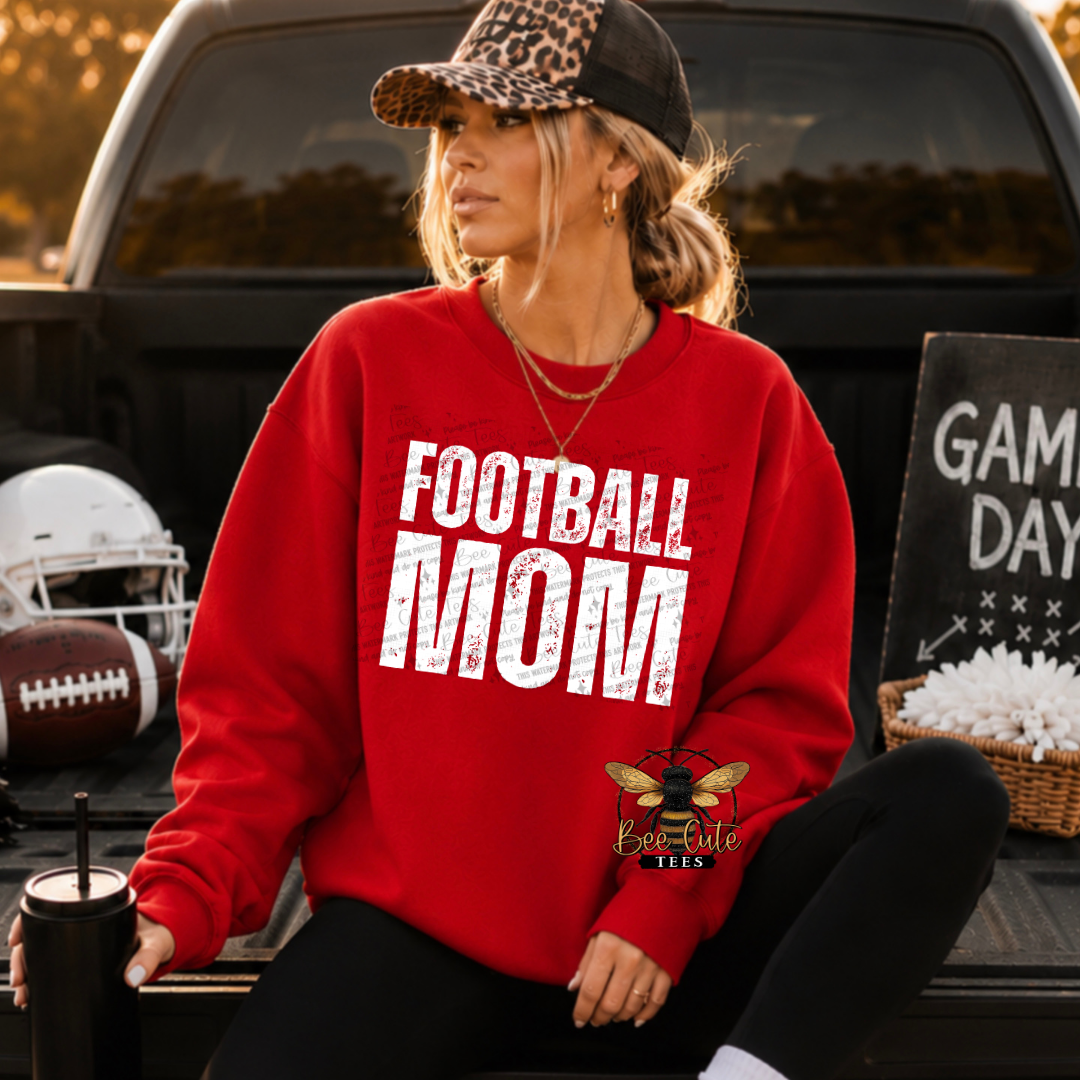 A woman sitting in the back of a truck wearing a red sweatshirt with 'FOOTBALL MOM' printed in white distressed letters. She is also wearing a black and leopard-print baseball cap and black pants.