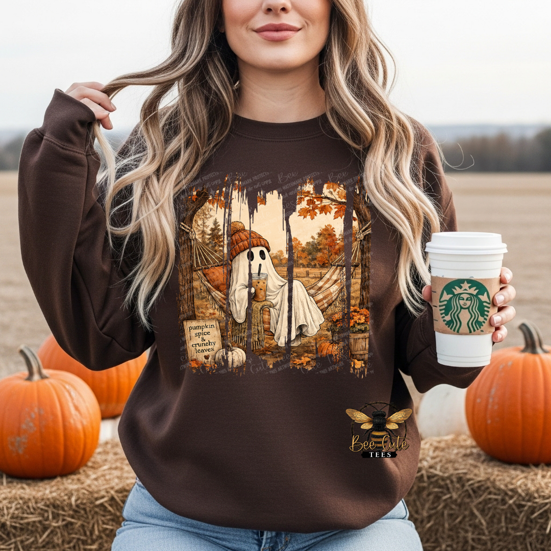 A woman wearing a brown crewneck sweatshirt with a graphic of a winged ghost in an autumn tree, holding a  coffee cup in a fall-themed outdoor setting with pumpkins and hay.