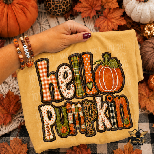 A folded mustard yellow T-shirt is held by a person's hand over a fall-themed background with pumpkins, leaves, and a plaid blanket. The shirt features a large applique-style 'hello pumpkin' design with multiple patterned letters and an orange pumpkin motif.