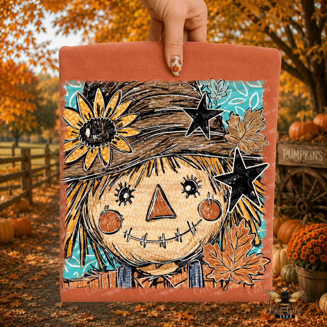 A hand holds a folded orange t-shirt with a whimsical scarecrow illustration with a sunflower, black stars, autumn leaves, and a turquoise background. The scene is set outdoors in a fall yard with pumpkins, orange foliage, and a wooden fence.