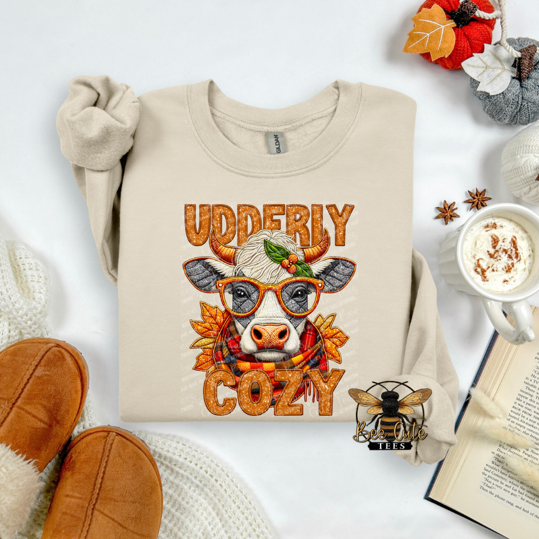 A beige crewneck sweatshirt laid flat with rolled sleeves, featuring a cow illustration wearing glasses and a scarf with the words 'UDDERIY COZY' in orange textured lettering. The sweatshirt is styled in a cozy autumn flat lay with slippers, a mug of hot cocoa, an open book, and fall leaves.