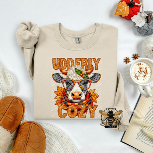 A beige crewneck sweatshirt laid flat with rolled sleeves, featuring a cow illustration wearing glasses and a scarf with the words 'UDDERIY COZY' in orange textured lettering. The sweatshirt is styled in a cozy autumn flat lay with slippers, a mug of hot cocoa, an open book, and fall leaves.