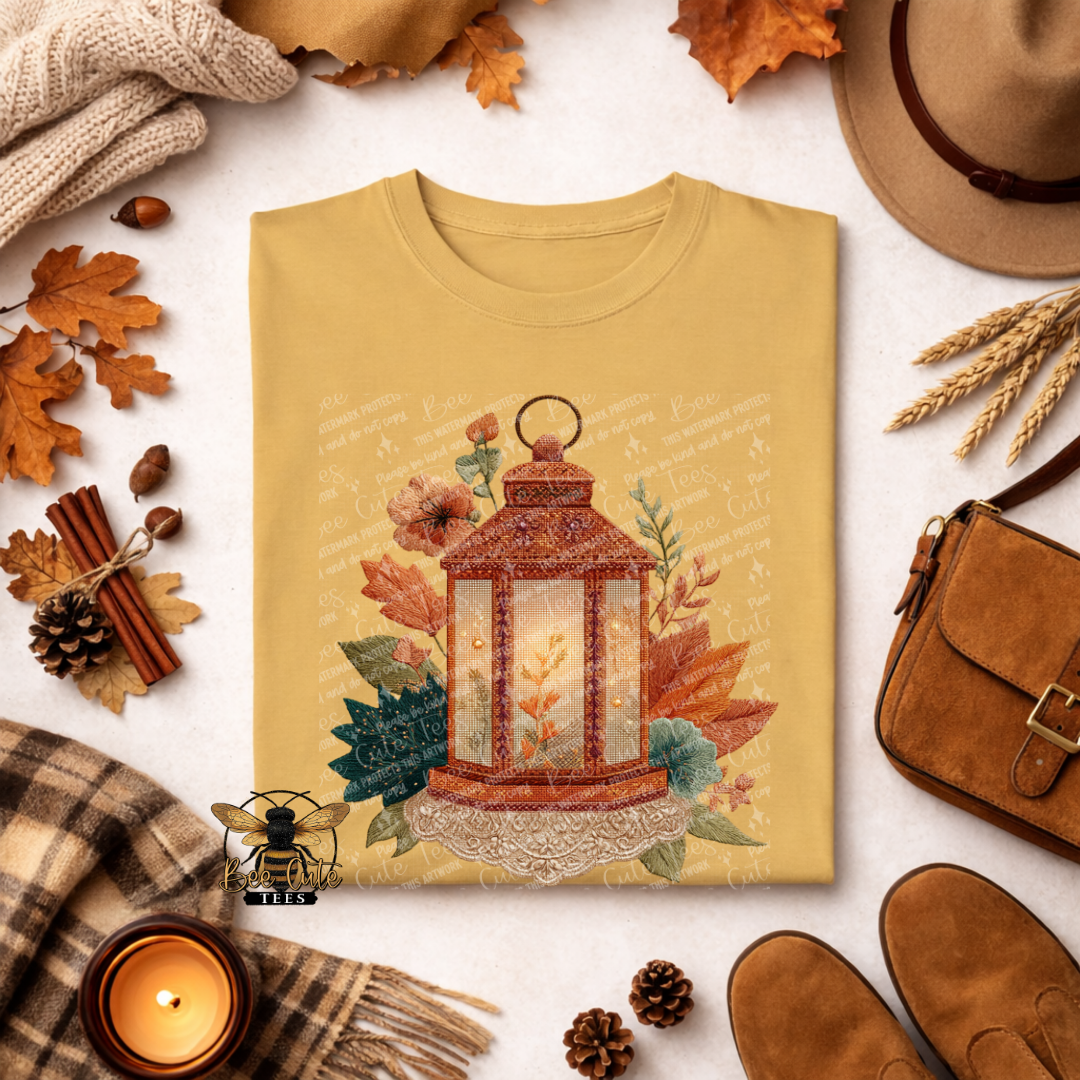Folded mustard yellow t-shirt featuring a vintage cottage-style lantern with a warm glowing center, surrounded by rust-colored fall leaves, delicate flowers, and sage green foliage. Intricate faux embroidery textures and cream lace accents complete the cozy autumn design. Styled with a candle, plaid blanket, dried leaves, and rustic accessories. Bee Cute Tees watermark visible.