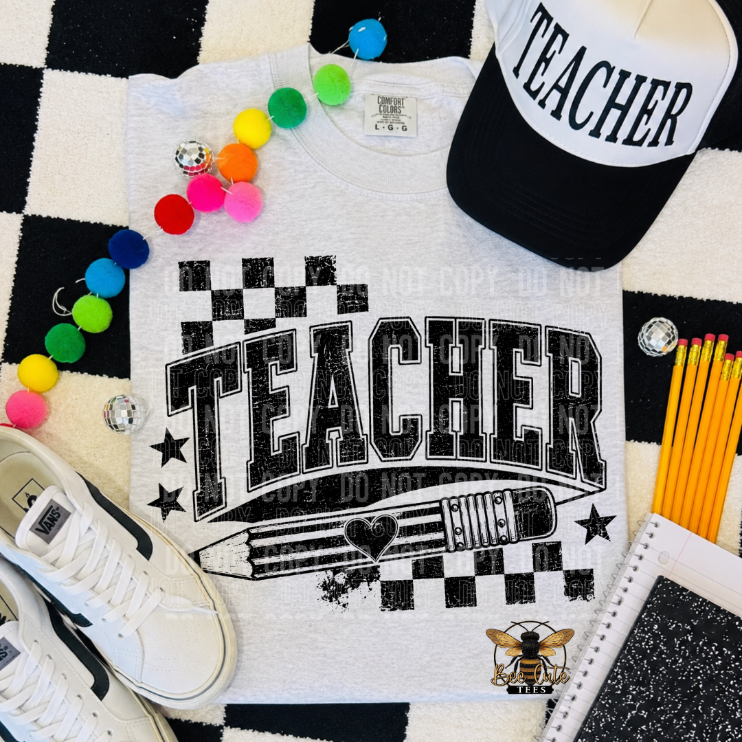 A white graphic T-shirt laid flat on a black-and-white checkered surface, surrounded by school-themed props including a black-and-white cap with 'TEACHER' text, colorful pom-poms, pencils, a notebook, and a shoe. The shirt has a large distressed 'TEACHER' print with stars, a pencil illustration, and a bee logo.