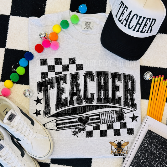 A white graphic T-shirt laid flat on a black-and-white checkered surface, surrounded by school-themed props including a black-and-white cap with 'TEACHER' text, colorful pom-poms, pencils, a notebook, and a shoe. The shirt has a large distressed 'TEACHER' print with stars, a pencil illustration, and a bee logo.