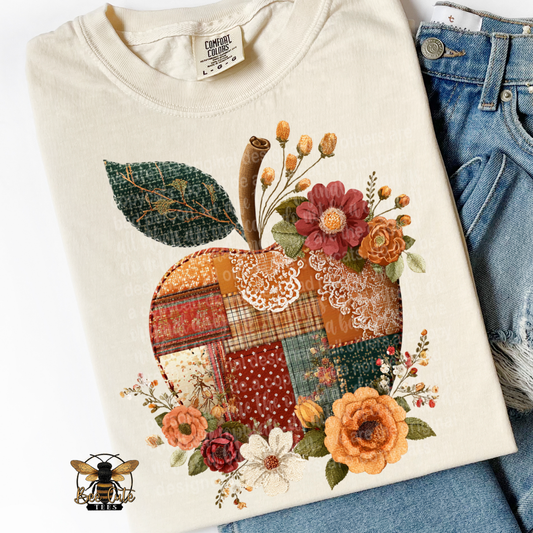 Folded ivory Comfort Colors t-shirt featuring a vintage cottage-style apple design with colorful faux patchwork fabrics in rust, orange, cream, burgundy, and forest green. Decorated with delicate lace accents, autumn flowers, and greenery. Styled with distressed blue jeans and a Bee Cute Tees watermark.