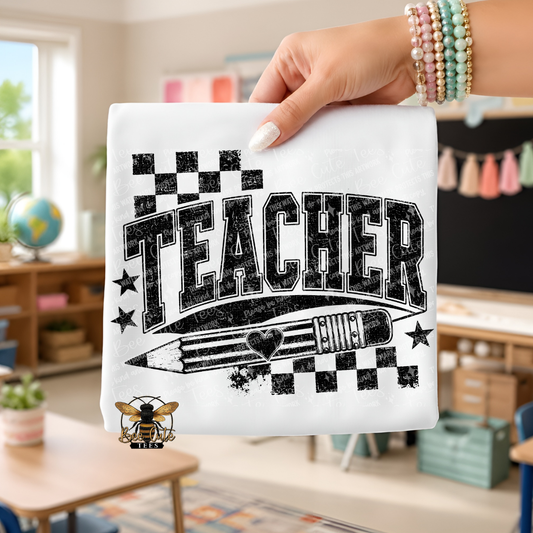 A hand holds a white folded t-shirt featuring a black distressed graphic that reads "TEACHER" with stars, checkered flags, and a pencil illustration. The background shows a blurred classroom interior with desks, a globe, and wall decorations.