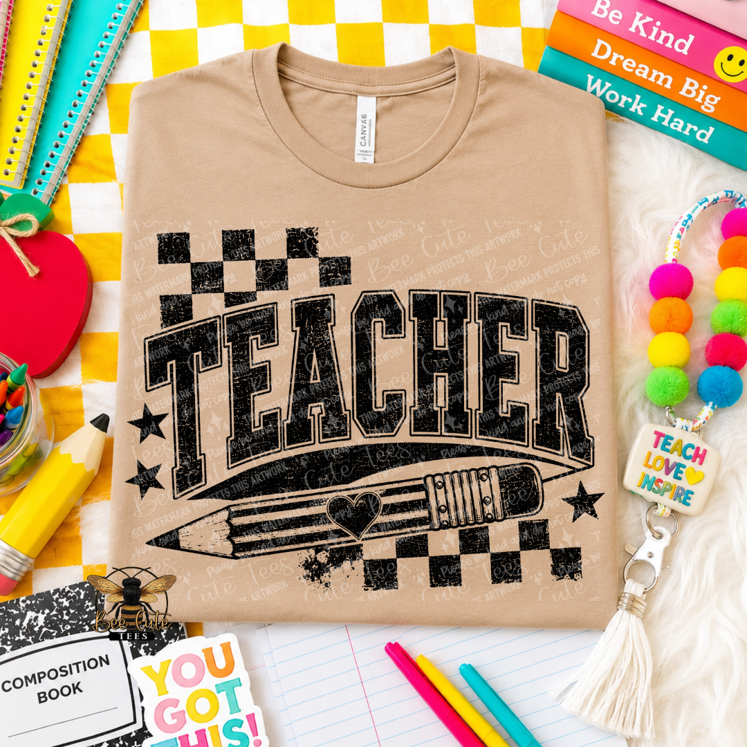 A beige graphic t-shirt featuring the word 'TEACHER' in distressed black block letters with a pencil and checkered design underneath. The shirt is displayed in a flat-lay arrangement with school supplies, books, and a colorful pom-pom keychain on a yellow-and-white checkered background.