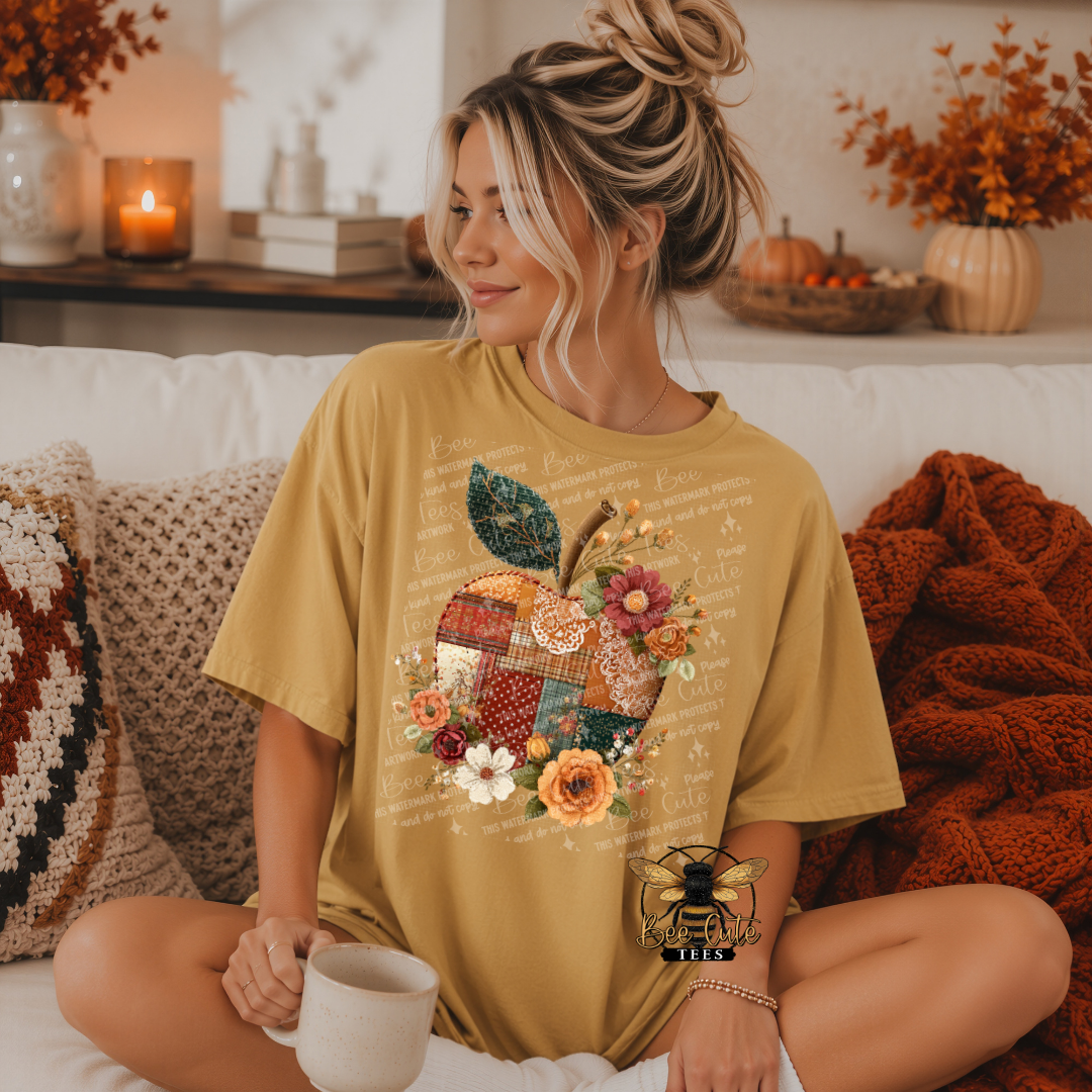 Woman wearing a mustard yellow t-shirt featuring a vintage patchwork apple decorated with floral patterns, faux embroidered textures, lace accents, and flowers in warm autumn colors. Relaxing on a white couch with a coffee mug, surrounded by cozy blankets, candles, and fall decorations. Bee Cute Tees watermark visible.