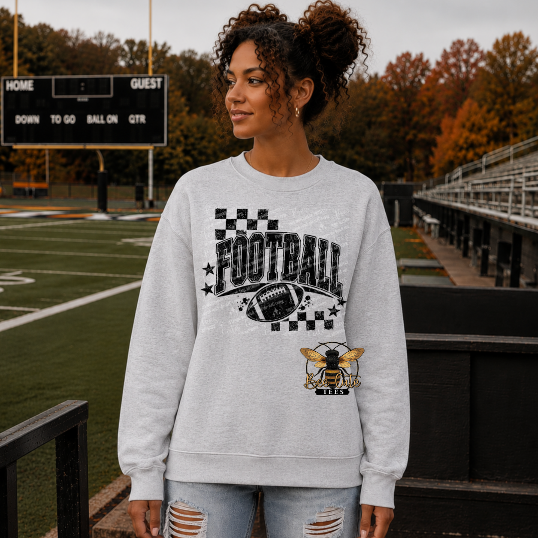 A woman stands on a football field wearing a light gray crewneck sweatshirt with a black and white 'FOOTBALL' graphic, a football, and a bee logo design. The background shows the green turf, bleachers, and autumn trees.