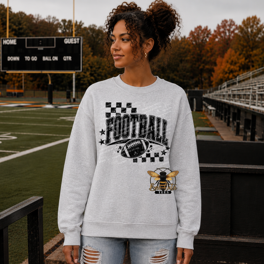 A woman stands on a football field wearing a light gray crewneck sweatshirt with a black and white 'FOOTBALL' graphic, a football, and a bee logo design. The background shows the green turf, bleachers, and autumn trees.