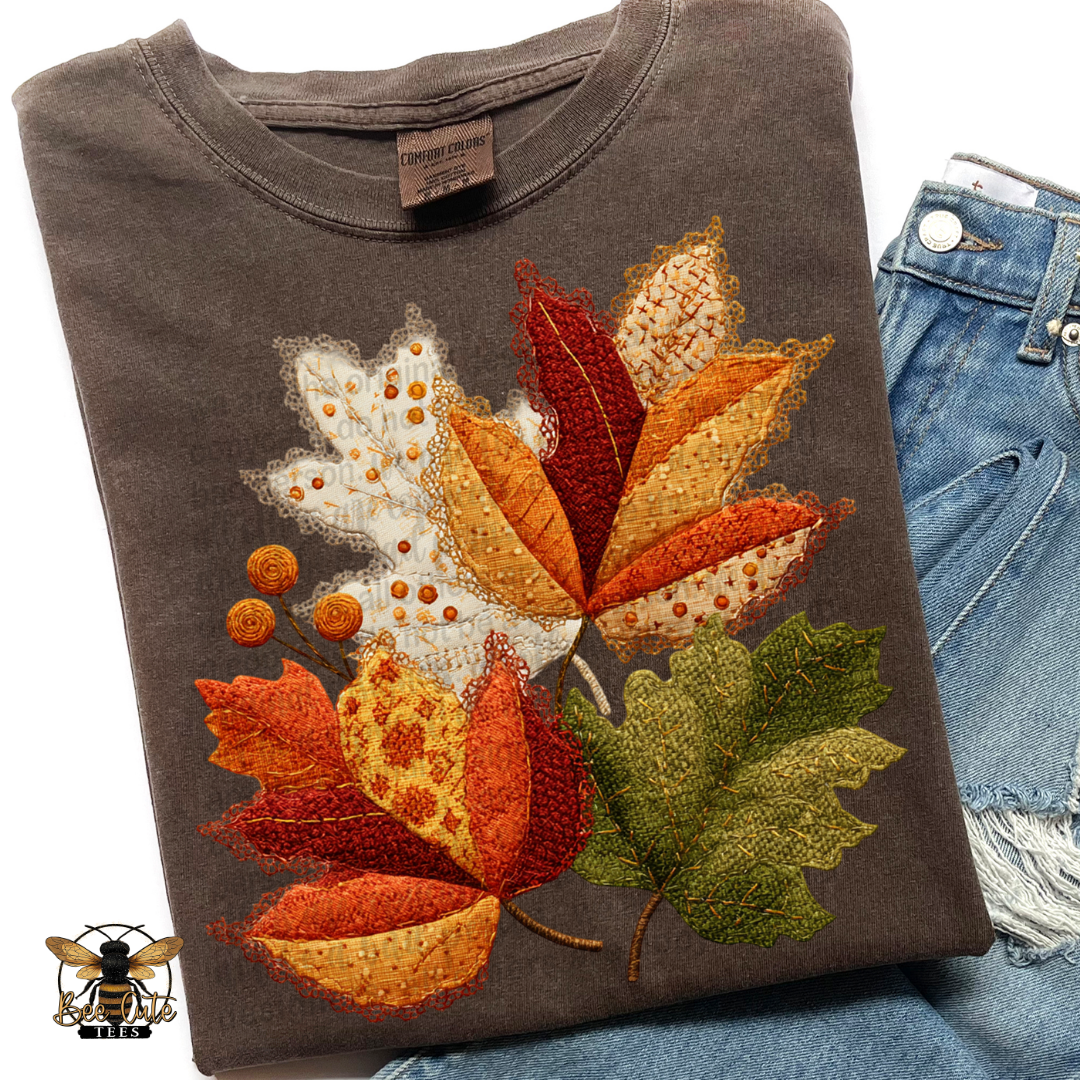 Folded vintage brown Comfort Colors t-shirt featuring a cottage-style arrangement of autumn leaves with faux embroidery, patchwork fabrics, and delicate lace textures. Rich shades of burnt orange, rust, burgundy, cream, mustard, and olive green. Styled with distressed blue jeans and a Bee Cute Tees watermark.